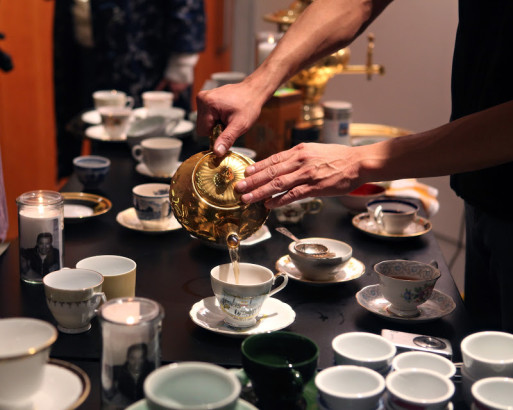 A pair of hands gently holds a tea pot over a table filled with delicate ceramic teacups.