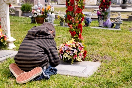 A grieving child kneels crying at flower wreath covered gravestone