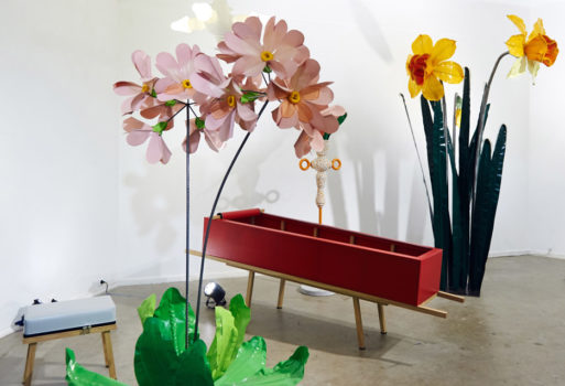 A red, zip-up coffin sitting next to two whimsical flower art pieces