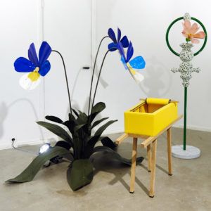 A yellow, zip-up coffin sitting next to a whimsical PVC plant statue
