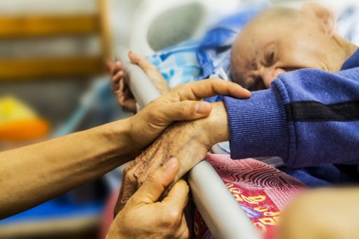 A hospice patient holds a volunteer's hand from a hospital bed moving towards a good death