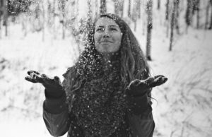 A woman stands in the snow with her arms out, smiling while snowflakes fall on her