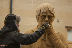 An artist finishes sculpting the sand on the woman's face, which looks much older now, with more wrinkles