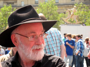Terry Pratchett stands outside near a crowd of people, wearing his signature reading glasses and black fedora