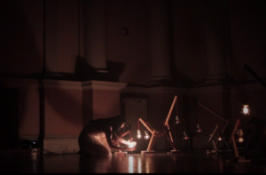 A dancer crouches next to three lightbulbs on the ground and creates shadows behind her, expressing one of the five stages of grief