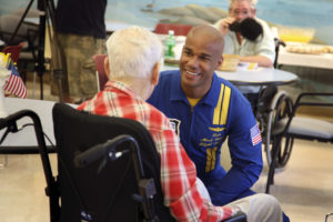 A man in a nursing home sits in a wheelchair and speaks with a member of the US Navy