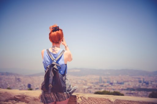 A woman wearing a backpack sits on the edge of a ledge, looking at the city below