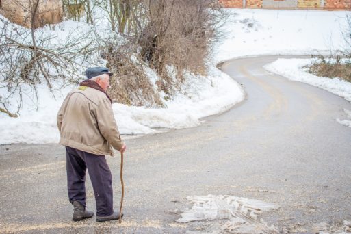 Elderly man walking down a windy road with a walking stick.