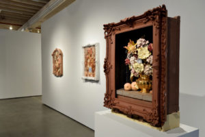 A photo of the still life vanitas gallery show, featuring a wooden frame with real sculpted objects inside of it, like a full flower bouquet