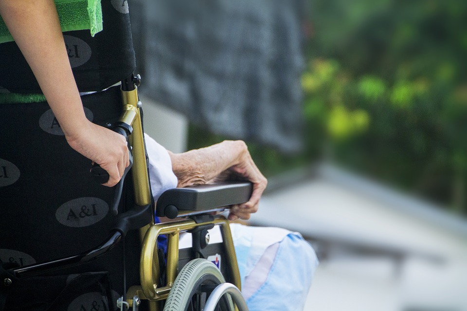 An elderly woman sits in a wheelchair, which is being pushed by a nursing home staff member