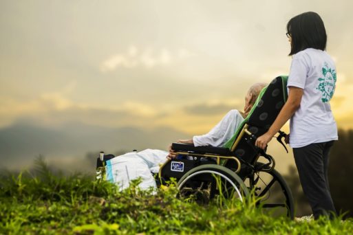 An elderly person is pushed in a wheelchair in a field of grass by a nurse, as she helps fulfill the last wishes of the dying