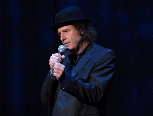 Comedian Steven Wright performing stand up with a microphone while wearing a dark suit and hat.
