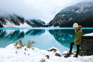 Woman standing next to a lake surrounded by fog and snow symbolizing travel related to a bucket list.