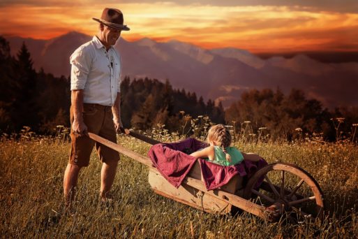 Father pushing daughter in a wheelbarrow through a meadow symbolizing life.