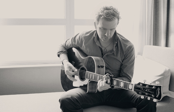 Black and white photo of musician Chris Trapper sitting on a couch and playing a guitar.