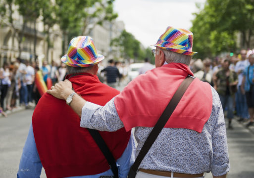 Two elderly men wearing rainbow-colored hats walking in a gay pride parade.