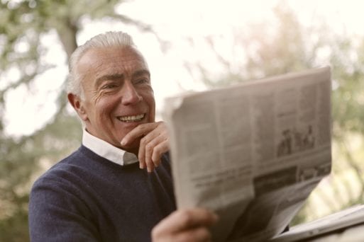 A man sitting outside laughing while reading a newspaper with funny obituaries
