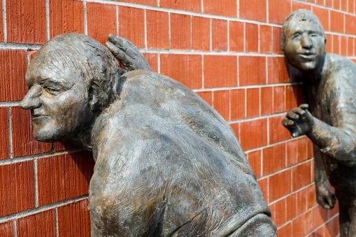 A sculpture of two elderly men with their ears to a brick wall demonstrating hearing and death