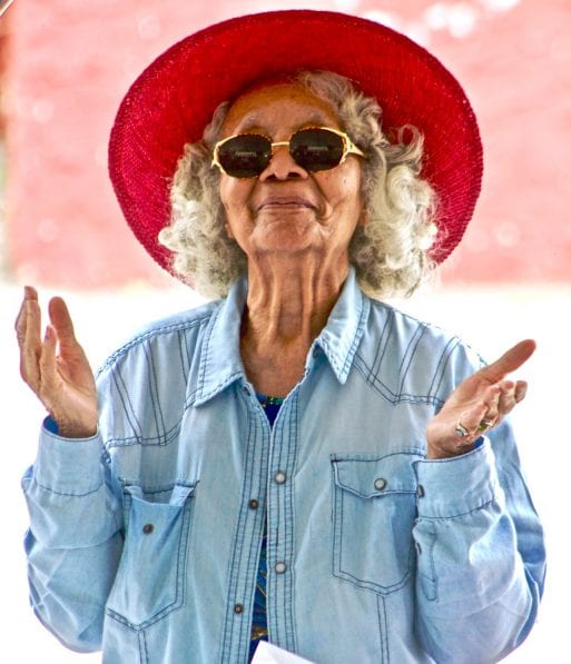 A senior woman in a bright red hat opens her hands in a happy stance.