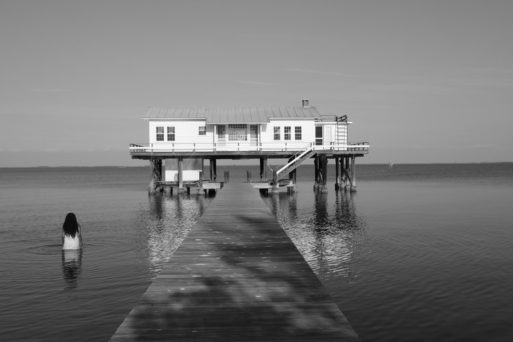 Black-and-white photo by Rachel Eliza Griffiths showing a woman standing in water next to a pier that leads to a small building