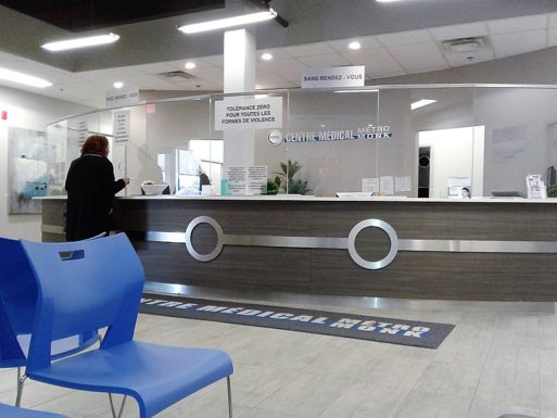 Hospital waiting room with one person standing at reception counter, one empty chair in foreground shows impact of non-COVID deaths.