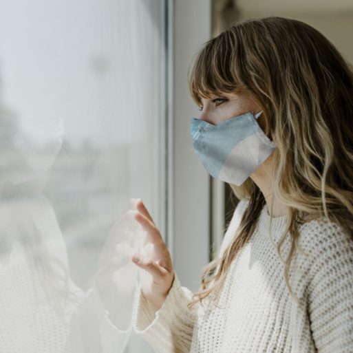 Blonde woman in beige sweater and face mask looking out of window with hand on glass (national grief strategy)