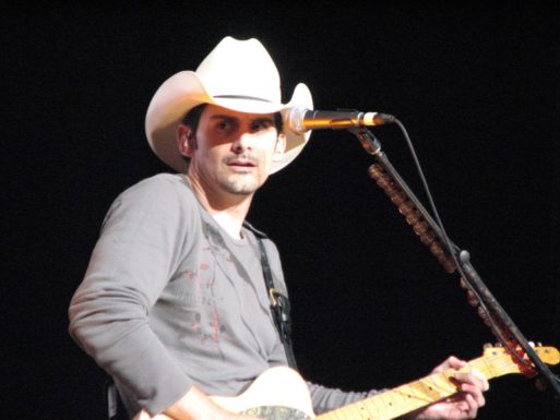 Brad Paisley in front of a microphone with a guitar, wearing a grey shirt and a cowboy hat with a black background - Whiskey Lullaby