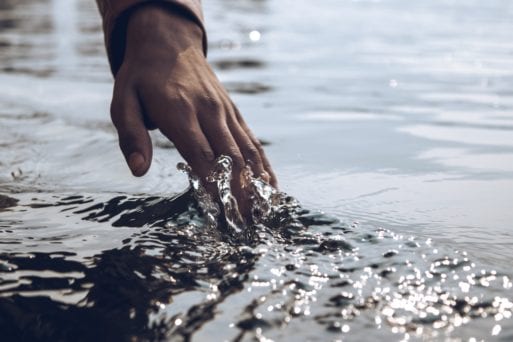 A hand touches water on a sandy shore.