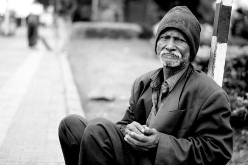 An elderly man in a beanie sits at the side of the road.
