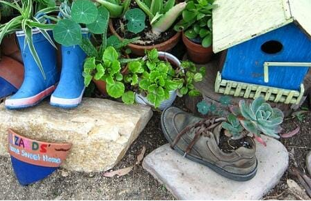 Blue boots and old shoes appear as planters amid pots, rocks and other garden items.