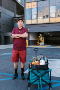 An elderly man stands next to a hand-drawn cart of groceries.