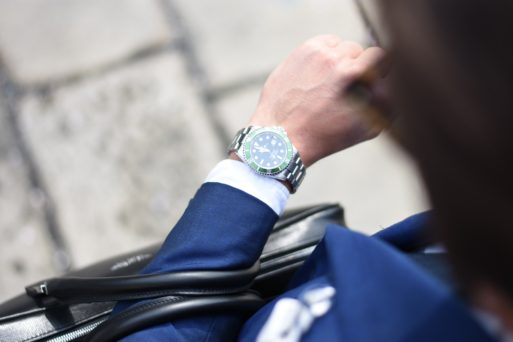 We look over a man's shoulder at his wristwatch; he's dressed in a blue suit with pocket square and the arm with the watch also holds a briefcase, so we can assume he's a businessman.