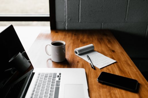 A laptop, cup of coffee (black), a cell phone, and a notebook with pen lie on a wooden table in front of a window.