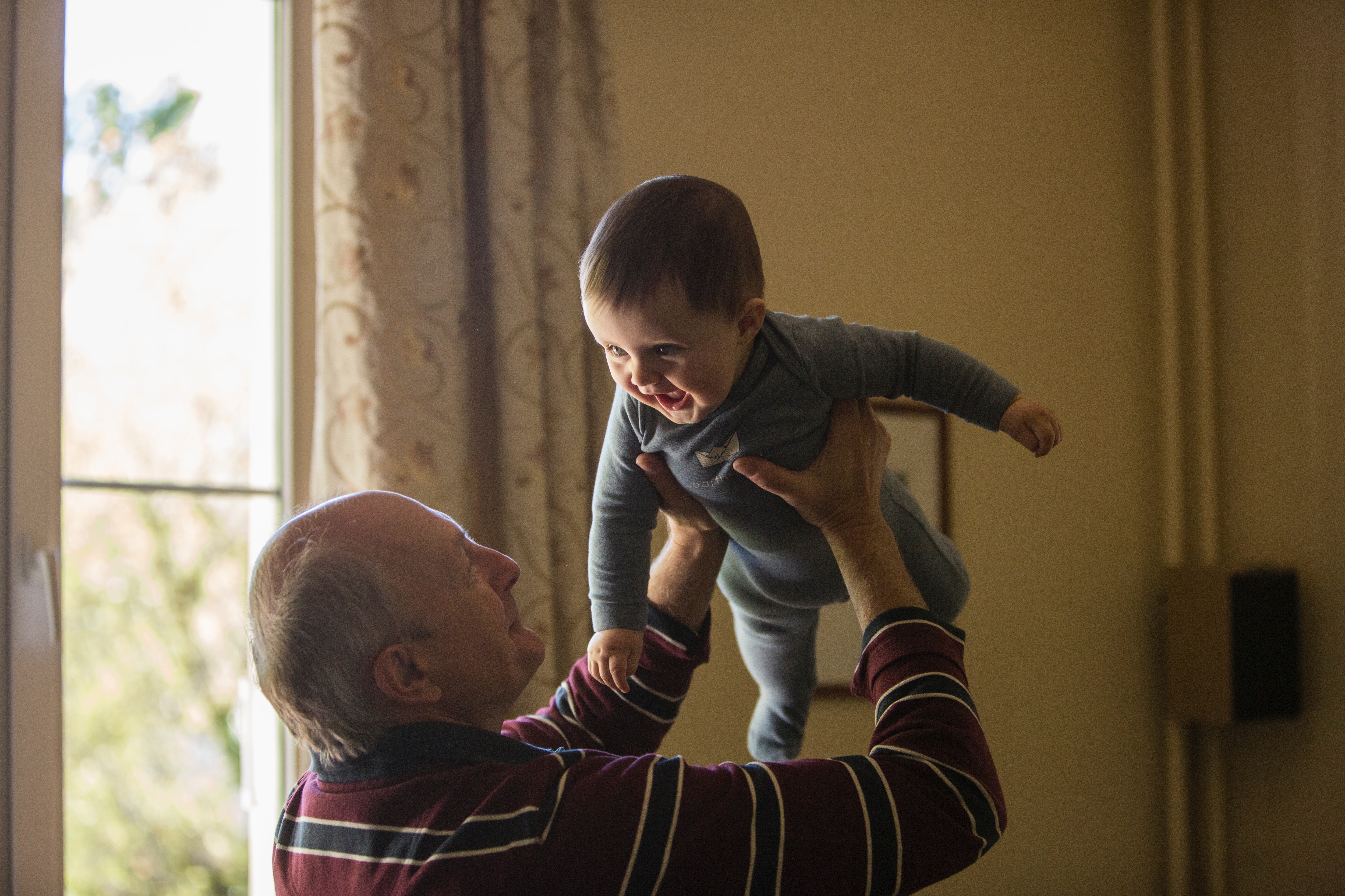 An older man swings a cute baby up in the air over his head, causing the baby to smile with glee. 
