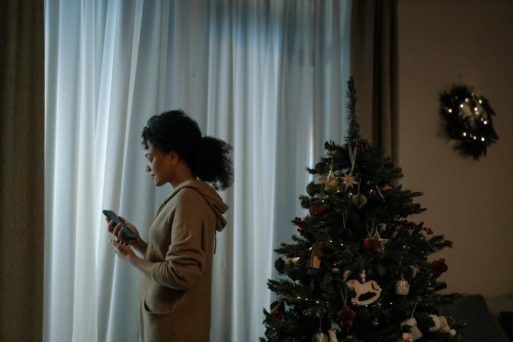 A woman looks at her phone in front of a Christmas tree, mourning a death during the holidays.