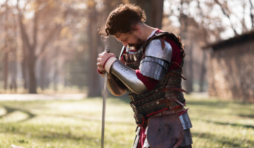A man in a medieval knight costume is kneeling in a field while grasping the pommel of his sword and bowing his head in grief, shows how Hollywood gets death wrong