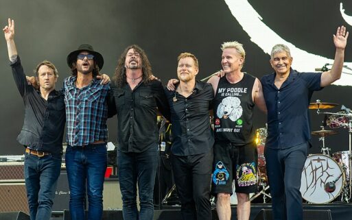 A picture of the Foo Fighters on stage with drummer Alex Frieze at the Glastonbury Festival in 2023. Six men in mostly black garb pose for a picture, with their arms around each other and smiling.