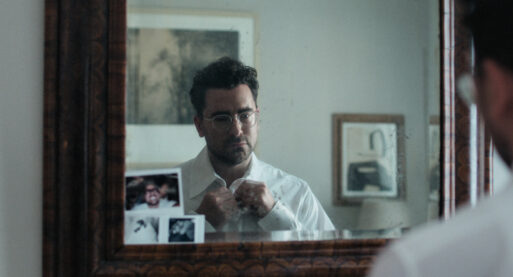 A man (actor Dan Levy) is sadly standing in front of a mirror but looking somewhere vaguely distant, buttoning up his white dress shirt. He is showing his grief at the loss of his husband.