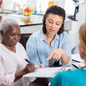A woman helps an elderly woman fill out medical paperwork while a nurse from the hospital looks on
