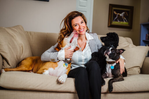 Dr. Dani McVety sits on a couch surrounded by three dogs. One is licking her chin while the other two gaze up at her. 