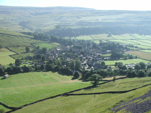 A panoramic photograph of Kettlewell in England shows a verdant, green expanse, rolling hills, and a small cluster of homes.