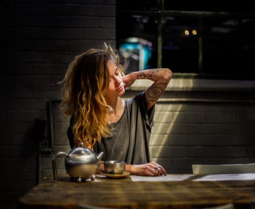 A woman sits at a table drinking tea in a dimly lit room. She's reaching her arm up as if to stretch out her stiff neck.