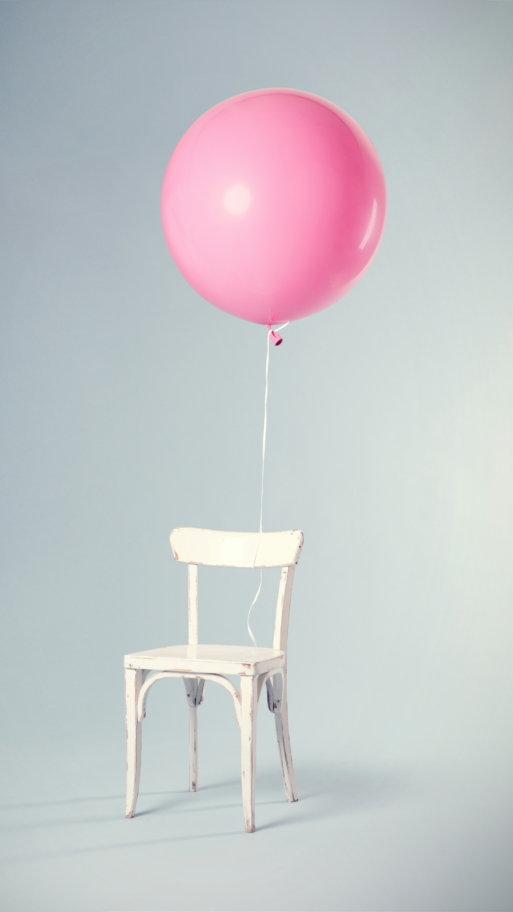 A white, wooden chair stands alone in a room with a single, pink, helium balloon tied to the back.