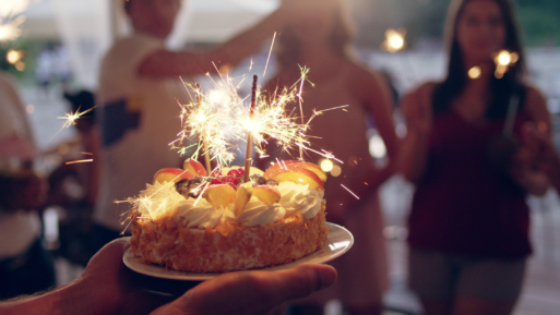 A beautifully decorated cake with lit sparklers as candles is being carried on a platter towards waiting guests. 