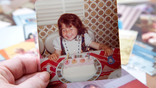 A hand is holding an old-looking photograph of a small girl smiling for the camera, sitting at a table with her birthday cake in front of her