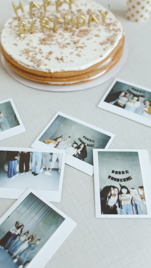 A birthday cake and scattered polaroid pictures sit on a white table, showing people at the party