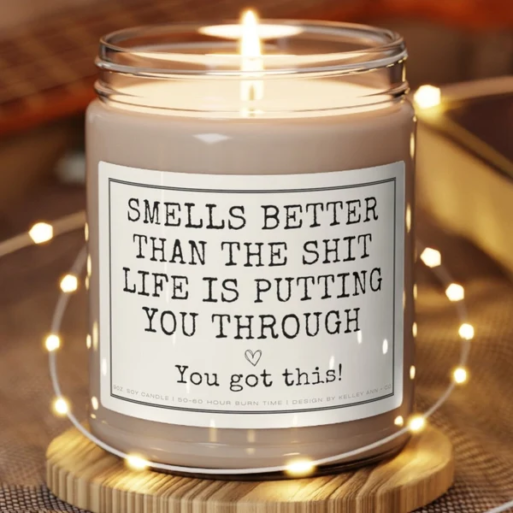 White candle in a glass jar, with a label that says, "Smells better than the shit life is putting you through; You got this!"