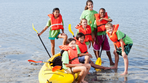 A group of kids wearing bright orange life jackets sit and stand around a yellow kayak in shallow water