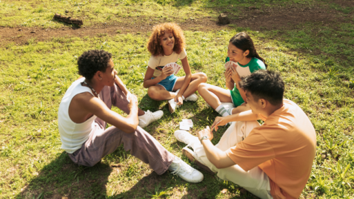 Four teenagers sit in a circle on a field of grass, playing a card game and smiling