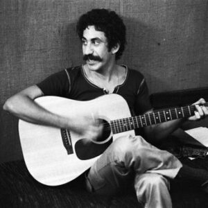 Jim Croce sitting against a wall, strumming his guitar